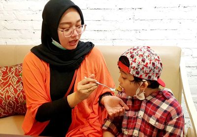 Mother feeding son while sitting at sofa