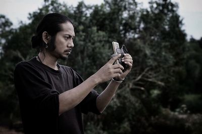 Man photographing with mobile phone while standing against trees