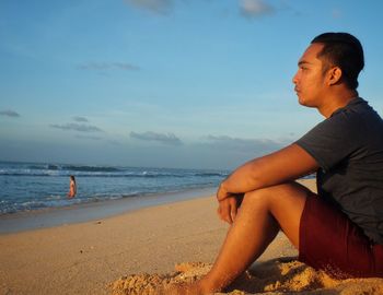 Side view of man sitting on beach against sky