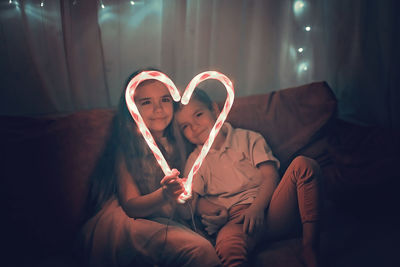 Siblings making heart shape of illuminated candy canes on sofa in darkroom