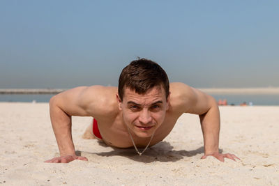 Portrait of shirtless man lying on sand at beach against sky