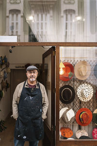 Content mature bearded male master in apron standing near showcase with collection of stylish handmade hats and looking at camera