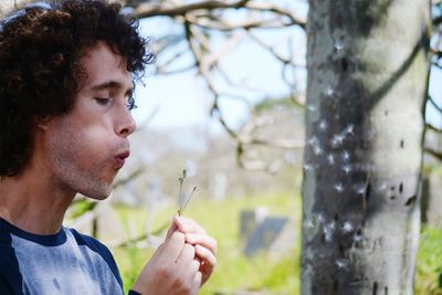 Close-up of man blowing dandelion against tree trunk