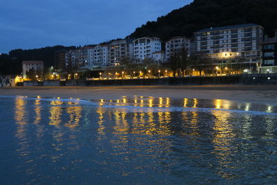 Illuminated buildings by lake against sky at night