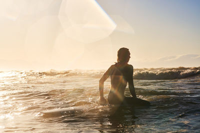 Young man with surfboard sitting on wavy sea at sunset