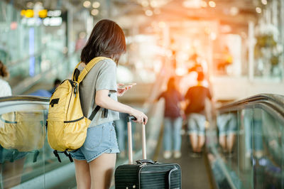 Rear view of woman with luggage using smart phone while standing on moving walkway