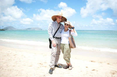 Full length portrait of happy mature couple standing at beach against sky during sunny day
