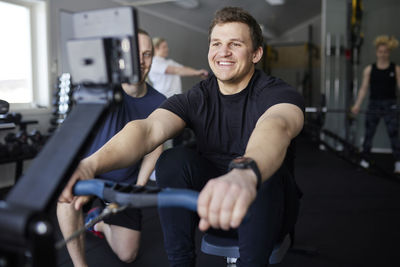 Smiling young man exercising on rowing machine by fitness instructor in gym