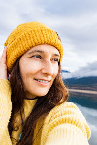 Woman with yellow sweater arranging her woolen hat to take a selfie with her mobile next to a lake