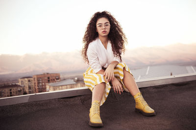 Portrait of young woman with curly hair sitting outdoors against sky