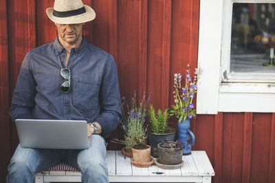 Man using laptop while sitting on bench by potted plants in back yard