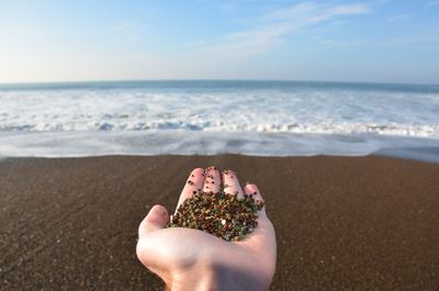 Cropped hand holding stones at beach against sky