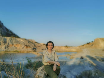 Portrait of smiling young woman sitting on rock against sky