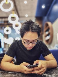 Young asian man in eyeglasses using smartphone at table in cafe against ceiling led lights.