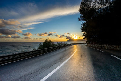 Road by trees against sky during sunset