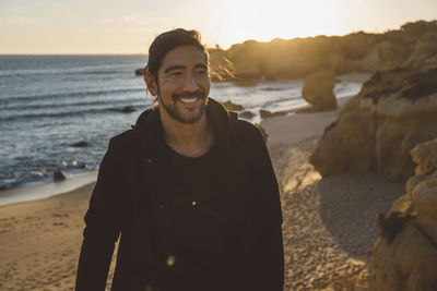 Smiling man looking away while standing at beach against sky during sunset