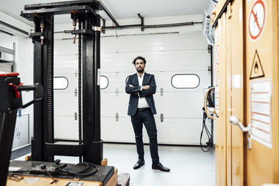 Confident businessman standing with arms crossed by forklift at industry