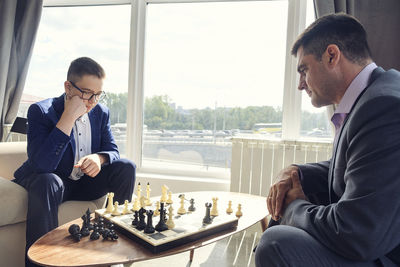 Dad and son are sitting at a table by a wide window and playing chess making moves with pieces