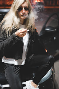 Portrait of young woman sitting on car,  woman at night wearing shades, sunglasses, bokeh