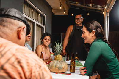 Cheerful diverse friends talking and smiling while sitting at table with beer bottles and vegetables sitting on terrace at night and having party