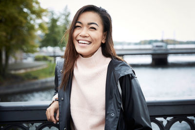 Portrait of happy woman wearing jacket standing on bridge in city