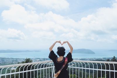 Rear view of woman making heart shape with arms against sea and sky