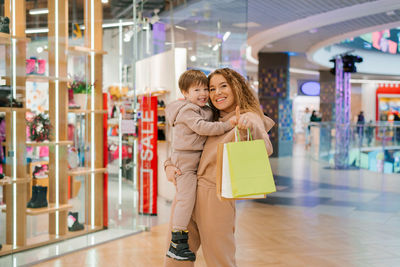 Smiling mother carries her little son in arms, holds shopping bags walks around the store on sale