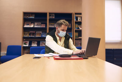 A young boy with a mask sitting and working with his laptop in the library