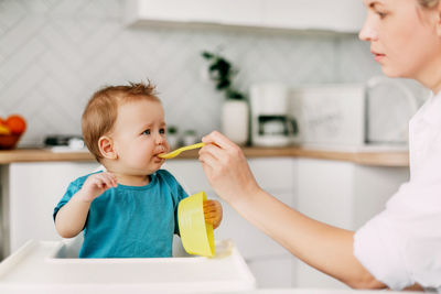 A young mother feeds her young son, who is sitting in a high baby feeding chair. care, motherhood, 