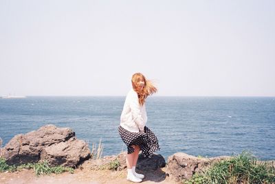 Full length of young woman standing on rock by sea against clear sky