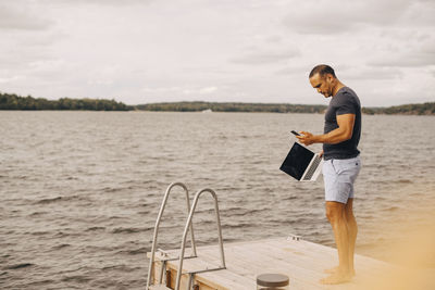 Full length of man with laptop using phone while standing on jetty against sky