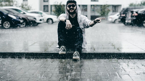 Portrait of young man gesturing while sitting on footpath during rain