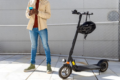 Unrecognizable african american male in casual style outfit standing on street near electric scooter and surfing internet on cellphone on sunny day