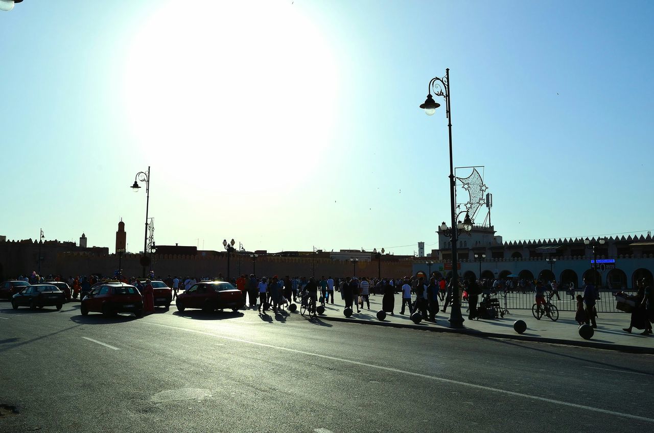 transportation, clear sky, land vehicle, street light, mode of transport, car, street, road, sunlight, blue, large group of people, sun, copy space, sky, men, city street, built structure, medium group of people, city