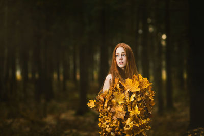 Thoughtful young woman covered with leaves standing at forest during autumn