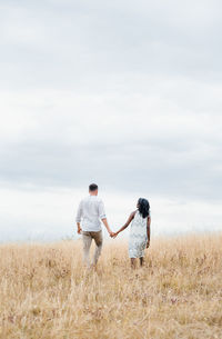 Back view of anonymous man with ethnic girlfriend holding hands while walking on autumn meadow under cloudy sky