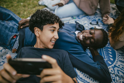 Smiling teenage boy holding smart phone and talking while lying on male friend