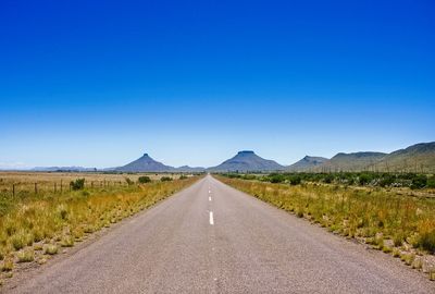 Road leading towards mountains against clear blue sky