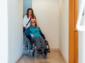 Content adult woman pushing wheelchair with senior mother and walking along corridor in rehabilitation center