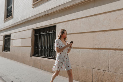 Unrecognizable female tourist in dress and mask text messaging on cellphone on pavement near stone building in seville