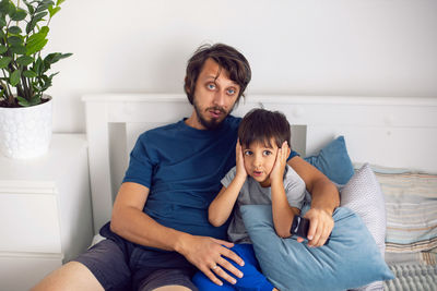 Bearded father and son are sitting on the bed in t-shirts and watching a football match on tv