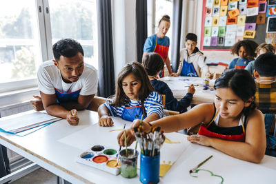 Art professor leaning on desk and teaching female pupils painting while sitting in classroom at school