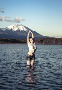 Young woman with arms raised standing in lake against snowcapped mountain