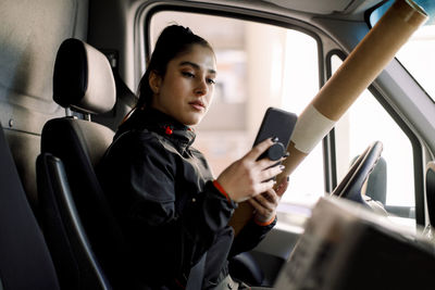 Young delivery woman using smart phone while holding package in truck
