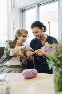 Senior woman teaching knitting to male nurse while sitting on sofa at elderly care home