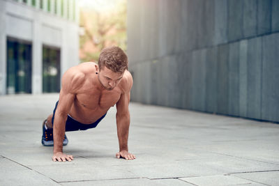 Full length of shirtless man doing push-ups on footpath by wall