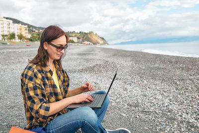 Woman in warm plaid shirt with laptop computer working outdoors while sitting on winter beach.