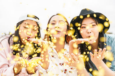 Portrait of happy friends blowing confetti while standing against sky