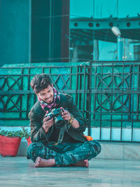 Full length of young man holding camera sitting outdoors