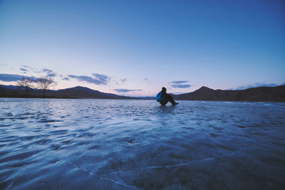 Silhouette mature man sitting on frozen lake against blue sky during sunset